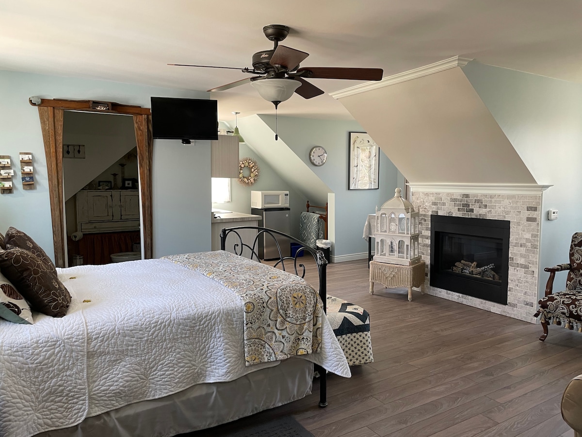 An open space showcases a queen-size bed with a patterned quilt and decorative pillows. A ceiling fan hangs above, while a television is mounted on the wall. A kitchenette is visible in the background, alongside a cozy sitting area featuring a decorative fireplace.