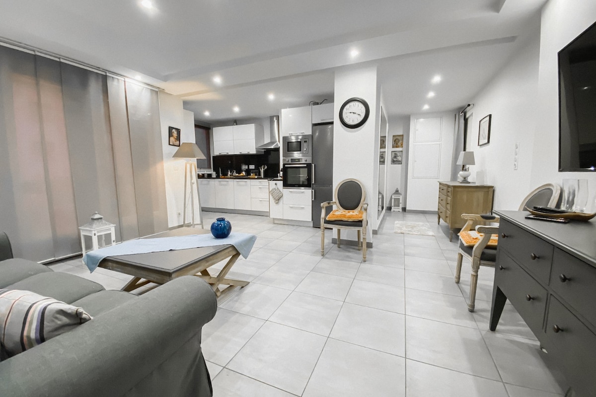 A modern living area is showcased with an open kitchen, featuring sleek cabinetry and contemporary appliances. Soft lighting enhances the spacious feel. A gray couch sits opposite a coffee table, while decorative elements, such as a lantern and vase, add subtle touches to the space.