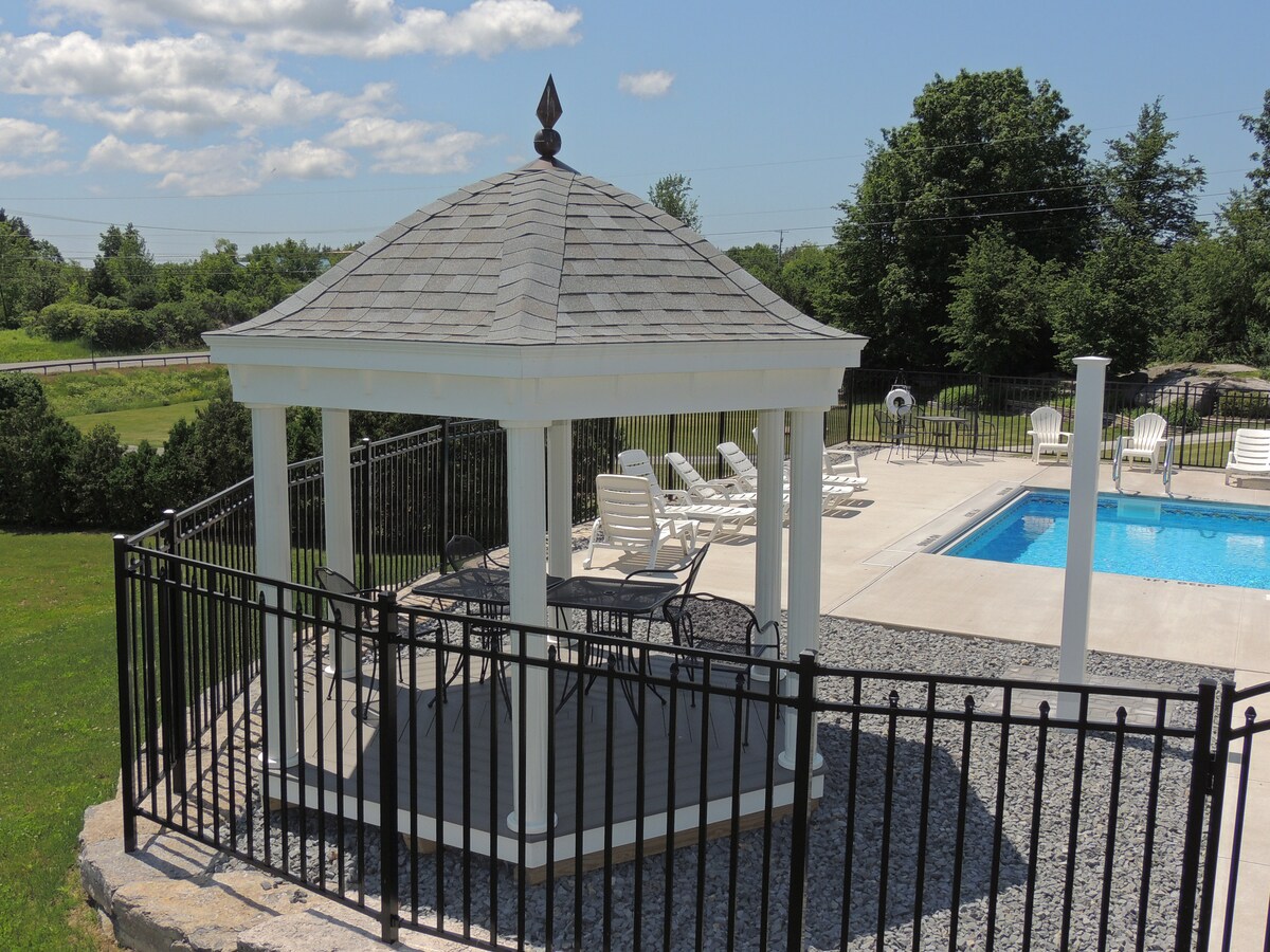 A gazebo with a shingled roof is seen near a seasonal heated saltwater pool. Surrounding the pool are several lounge chairs positioned on a concrete deck, while a black fence encloses the area. Lush greenery is visible in the background under a clear blue sky.