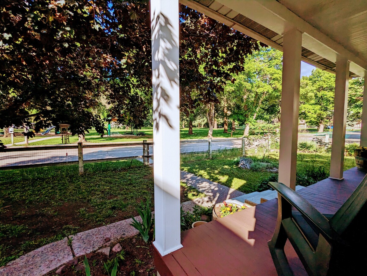 The image captures a serene porch area, showcasing wooden flooring and comfortable seating. Lush greenery can be seen in the foreground, with shade provided by large trees. A view of the quiet street and nearby park is visible, enhancing the peaceful atmosphere.