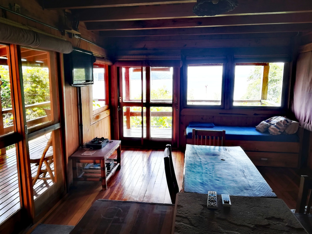 A wooden interior space features large windows that provide natural light and scenic views. A dining table is present, accompanied by chairs. A seating area includes a sofa, and a small television is mounted on the wall. The wooden floor adds a warm touch to the ambiance.