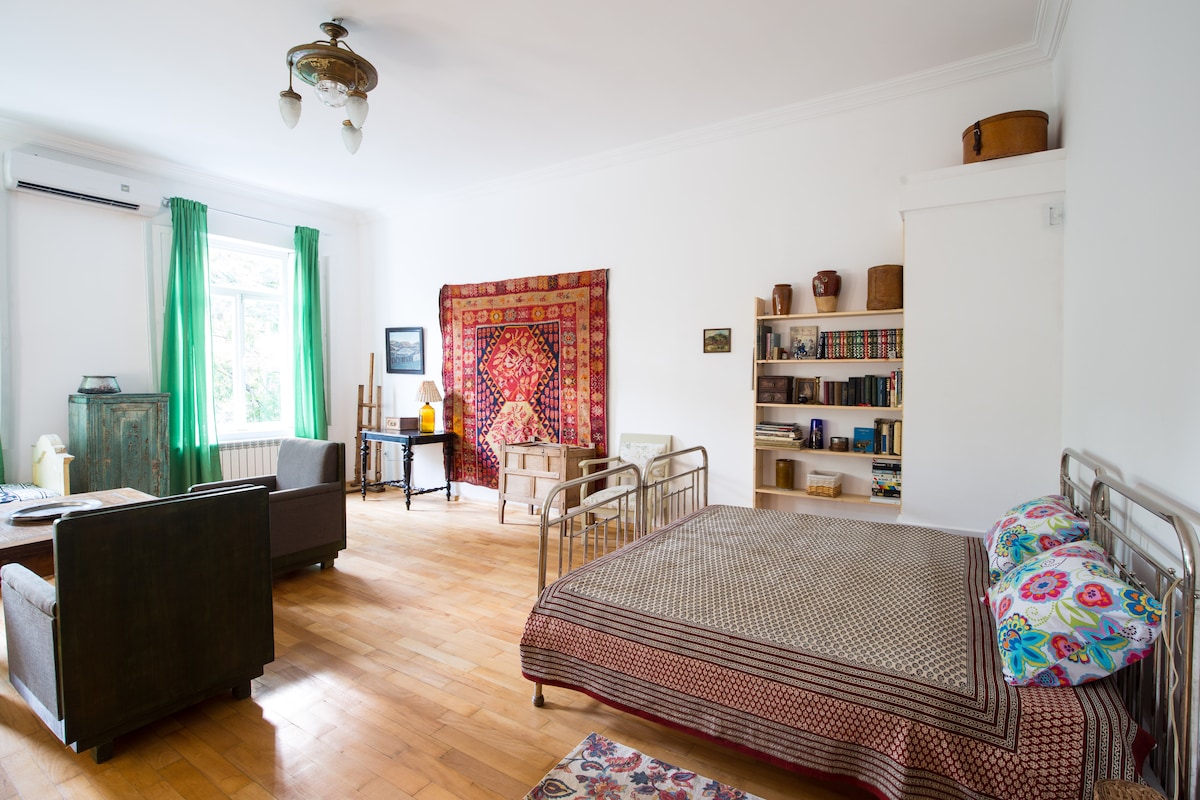 A spacious room features vintage-style furniture, including a large bed with patterned bedding. A decorative textile hangs on the wall, complemented by bookshelves filled with books. Natural light enters through large windows, with green curtains framing the view. A sitting area with two chairs is present.
