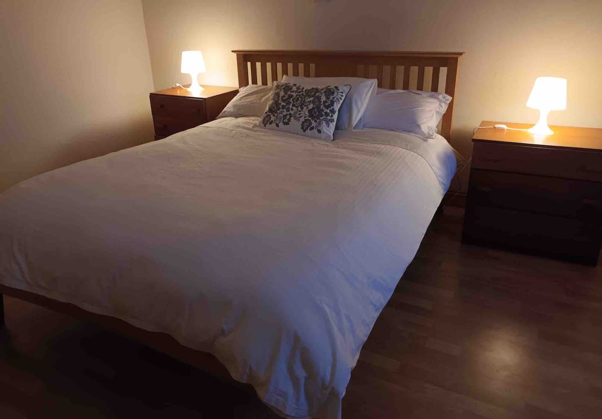 A spacious bed is positioned centrally in the room, complemented by two nightstands on either side. Each nightstand features a lamp, providing soft lighting that enhances the calming atmosphere. The room's wooden flooring contributes to a natural aesthetic.