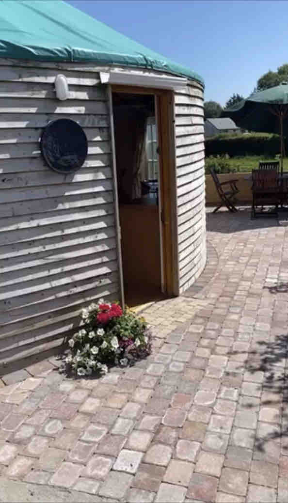 The entrance to Halfpenny Hut is framed by a circular wooden structure, featuring a welcoming door. A decorative plaque is mounted beside the entrance. A paved pathway leads to the door, where a flower arrangement adds a touch of color to the outdoor space.
