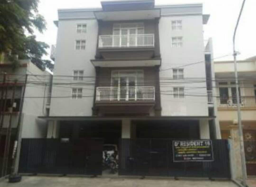 A modern three-story building features a minimalist facade with a blend of light and dark tones. The upper levels include balconies with railings, while the ground floor presents an open parking space. A large sign indicates the entrance to Kost Exclusive D'Resident 16.