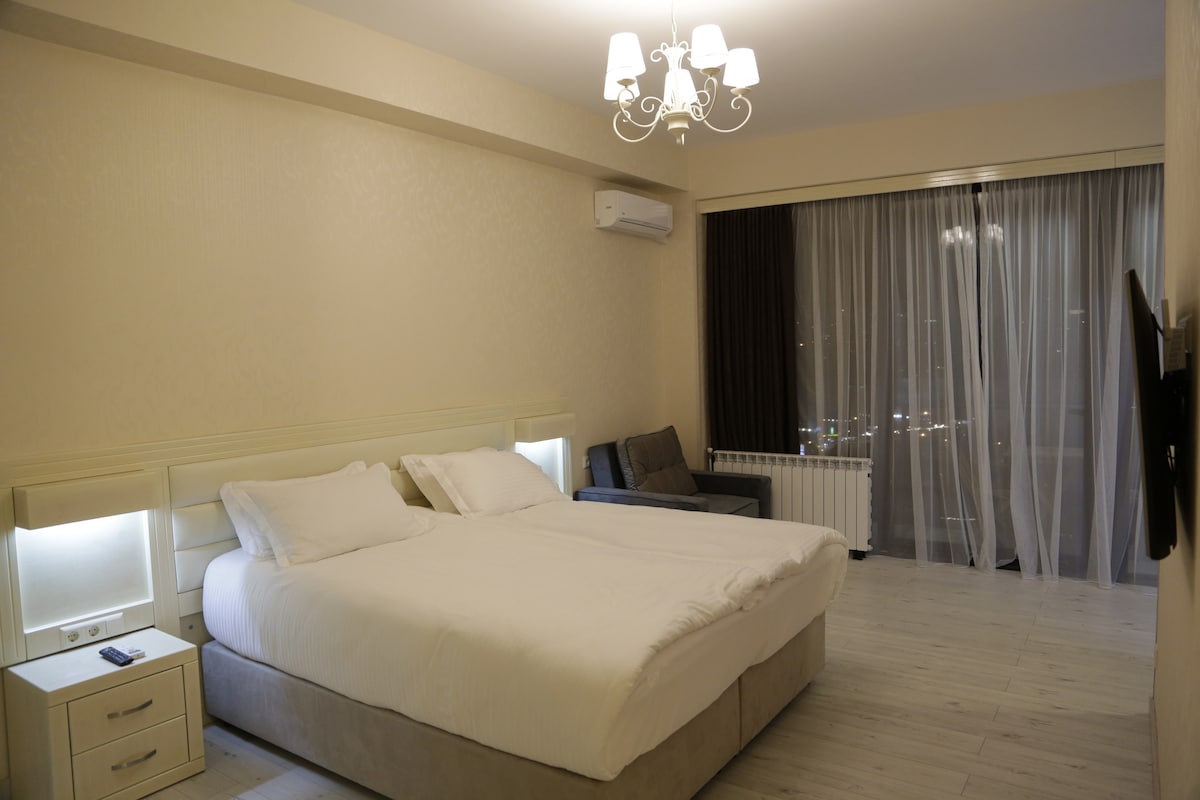 A spacious bedroom is presented with a large bed and a comfortable seating area. Soft lighting is provided by a chandelier, while sheer curtains frame a view of the city. A flat-screen TV is mounted on the wall, enhancing the room's functionality.
