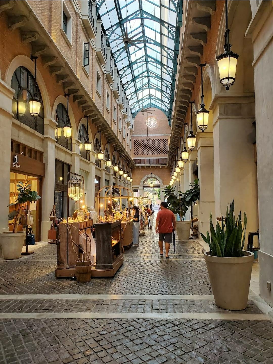 An indoor shopping area features a high glass ceiling allowing natural light to illuminate the space. Brick walls surround a cobblestone pathway, lined with potted plants and charming lighting fixtures. Various storefronts are visible, contributing to a lively atmosphere.