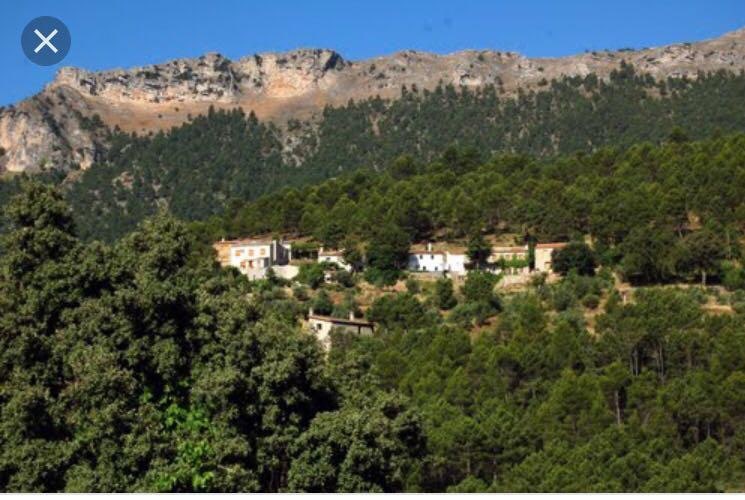 A rural setting is displayed, featuring several whitewashed buildings nestled among lush green trees, framed by majestic mountainous terrain. The clear blue sky enhances the tranquility of the scene, creating a serene atmosphere ideal for nature enthusiasts.
