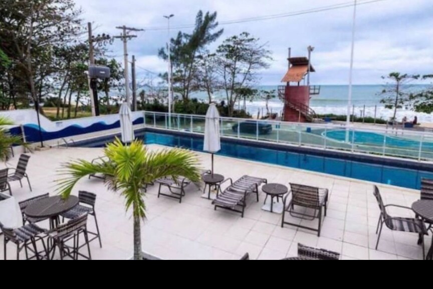 An outdoor pool area is visible, featuring a swimming pool with clear water surrounded by lounge chairs. A waterslide structure is located at one end, and palm trees add a touch of greenery. The ocean and sky with scattered clouds are seen in the background.