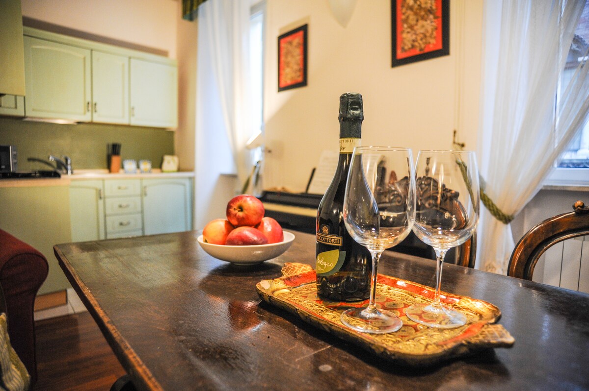 A dining area features a wooden table set with two glasses and a bottle of sparkling wine on a decorative tray. A bowl of fresh apples rests nearby. The modern kitchenette is visible in the background, illuminated by natural light filtering through sheer curtains.