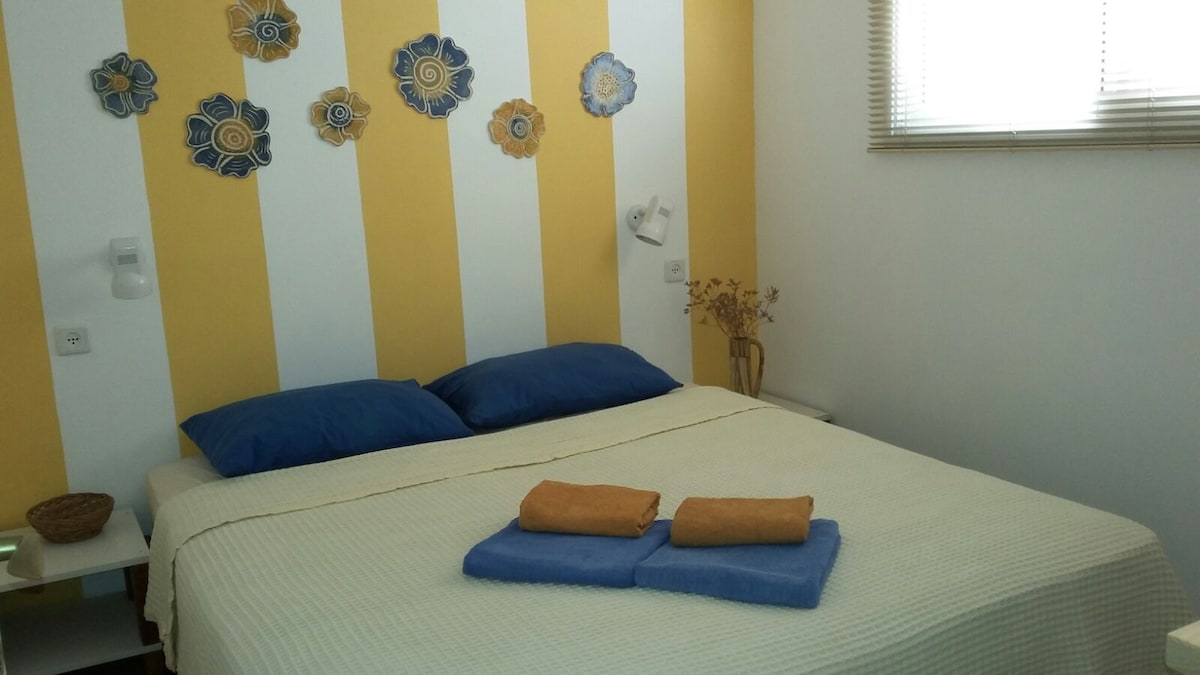 A cozy bedroom is presented, featuring a spacious bed with blue pillows and a neutral-toned bedspread. Soft towels are neatly arranged at the foot of the bed. The wall is adorned with decorative floral elements, and natural light is filtered through a window with blinds.