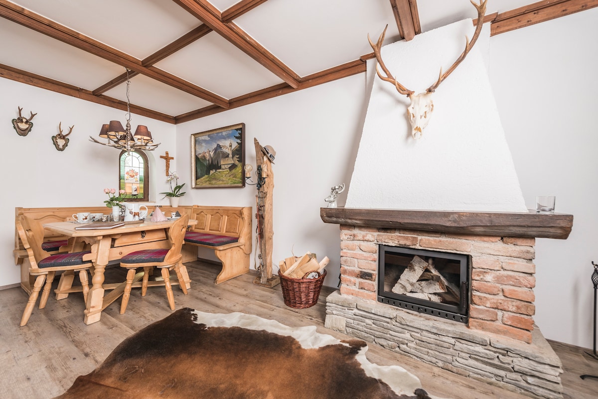 A cozy living area features a brick fireplace with a modern wood insert, surrounded by light wooden walls and a wooden ceiling with exposed beams. The dining table is set with decor and surrounded by comfortable seating. Antlers adorn the wall alongside rustic artwork.