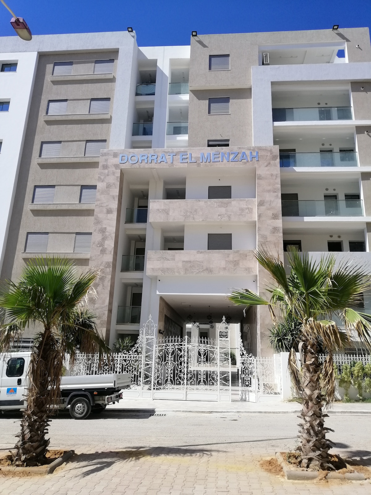 The entrance of the Dorrat El Menzah residence is depicted, showcasing a modern facade accented by palm trees. A security gate is visible, along with a welcoming lobby area. The building features a mix of concrete and glass elements, contributing to its contemporary design.