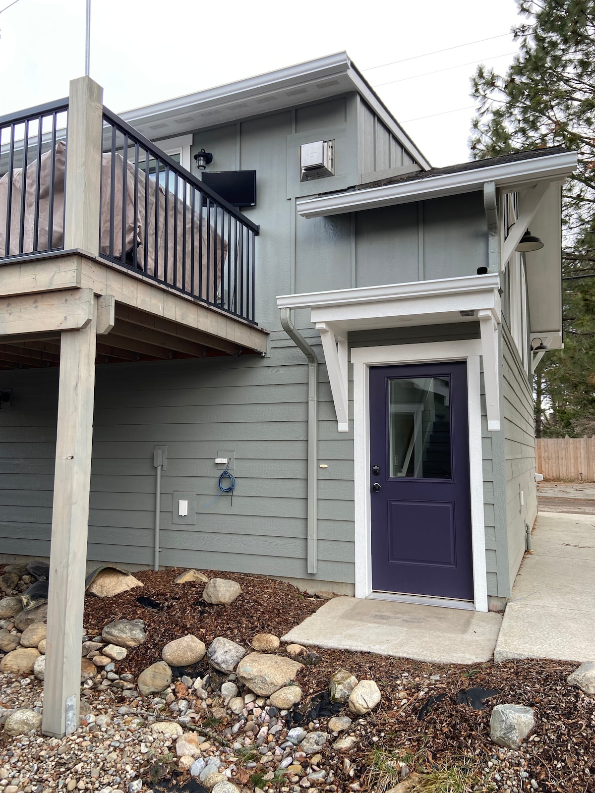 A modern entrance to the guest house features a deep purple door complemented by white trim. The building's light grey siding is accented by a spacious upper balcony. Natural stones are arranged along the walkway, leading up to the property.