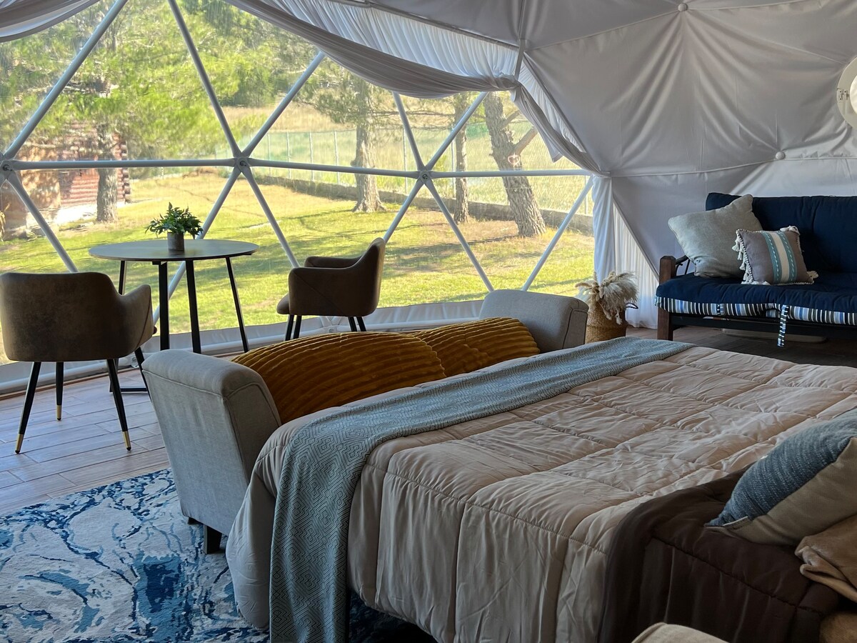 A cozy interior space features a spacious bed with a light-colored quilt, complemented by decorative pillows. A small table with two chairs is positioned nearby. Large windows offer a clear view of the natural surroundings, enhancing the connection to the outdoors.