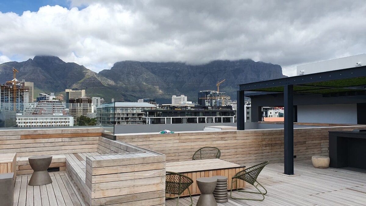 An outdoor rooftop space features wooden decking and seating arrangements. Tables and chairs in earthy tones are situated beneath a partly cloudy sky that highlights the distant Table Mountain, which serves as a dramatic backdrop to the stylish furnishings.