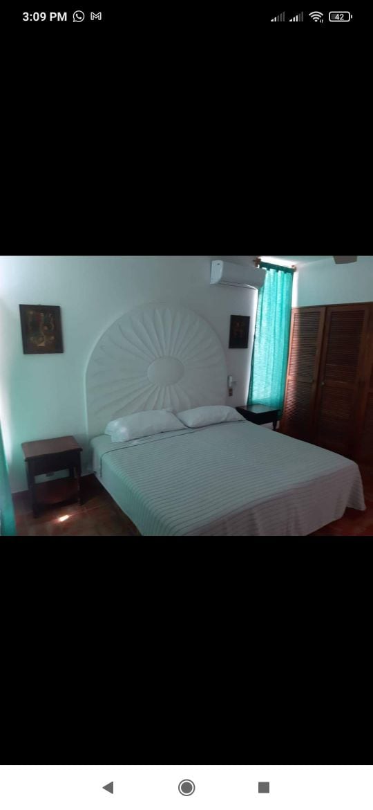 A well-appointed bedroom features a spacious bed with a striped blanket and decorative headboard. Natural light filters through turquoise curtains, illuminating the room. Two bedside tables are positioned on either side of the bed, and a wooden wardrobe is visible against the wall.
