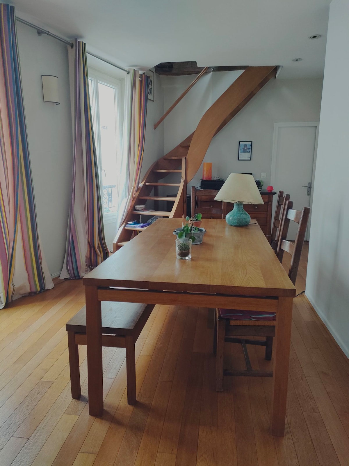 A wooden dining table accompanied by matching chairs occupies a well-lit space. Colorful striped curtains frame a window, while a small potted plant and a lamp add subtle touches to the table. A staircase is visible in the background, leading to the upper level.