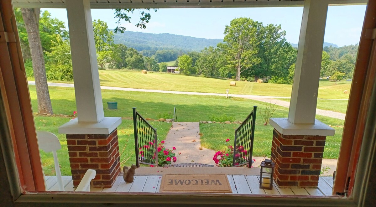 A scenic view from the front porch showcases rolling hills and lush greenery. The path leading to the porch is lined with vibrant flowers, while the inviting welcome mat rests on the wooden floor. Mountains can be seen in the distance, creating a serene backdrop.