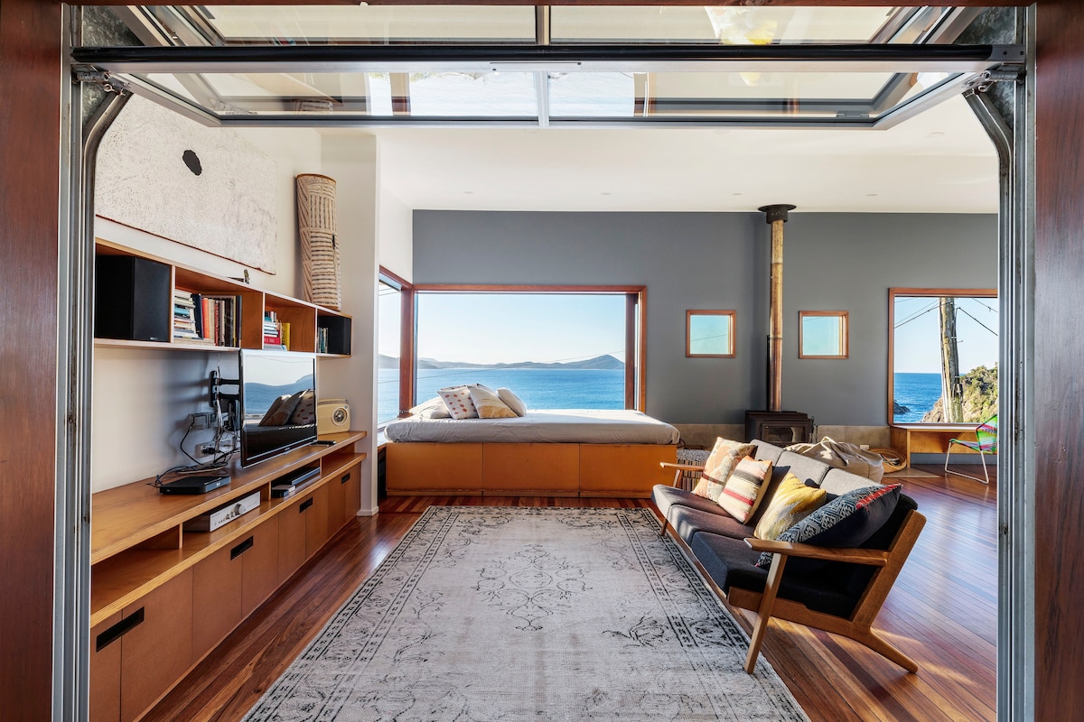 The open living area features large automated glass doors leading to a stunning ocean view. A cozy seating arrangement with a patterned rug complements a wooden media unit and a combustion fireplace, while a spacious bed is positioned near the window, offering an inviting, tranquil setting.