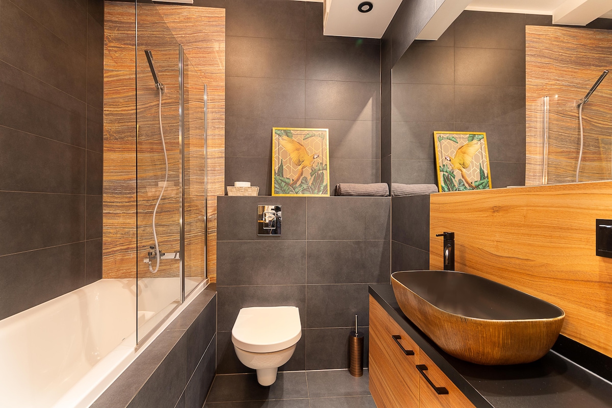 The modern bathroom features a combination of sleek dark tiles and warm wood accents. A spacious bathtub is complemented by a glass shower enclosure. A sculptural sink is positioned atop a wooden vanity, while two framed art pieces adorn the wall above.