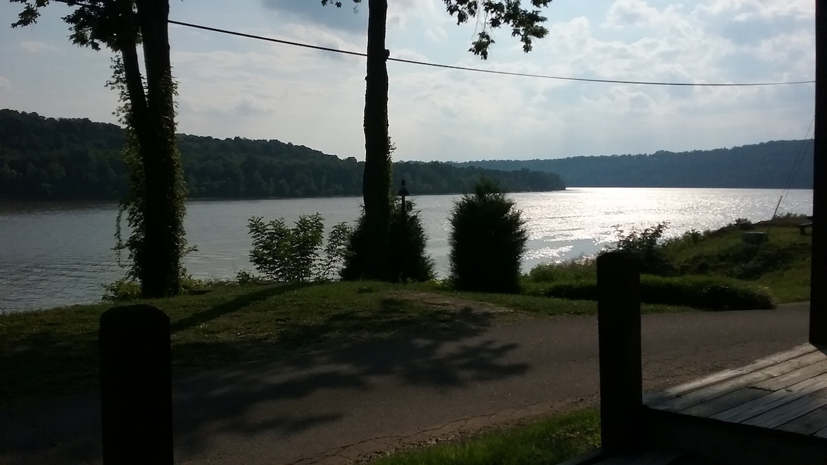 A serene view of the lake is framed by tall trees, with gentle sunlight reflecting off the water's surface. The shoreline features lush greenery, while rolling hills create a distant backdrop, offering a sense of peaceful seclusion.