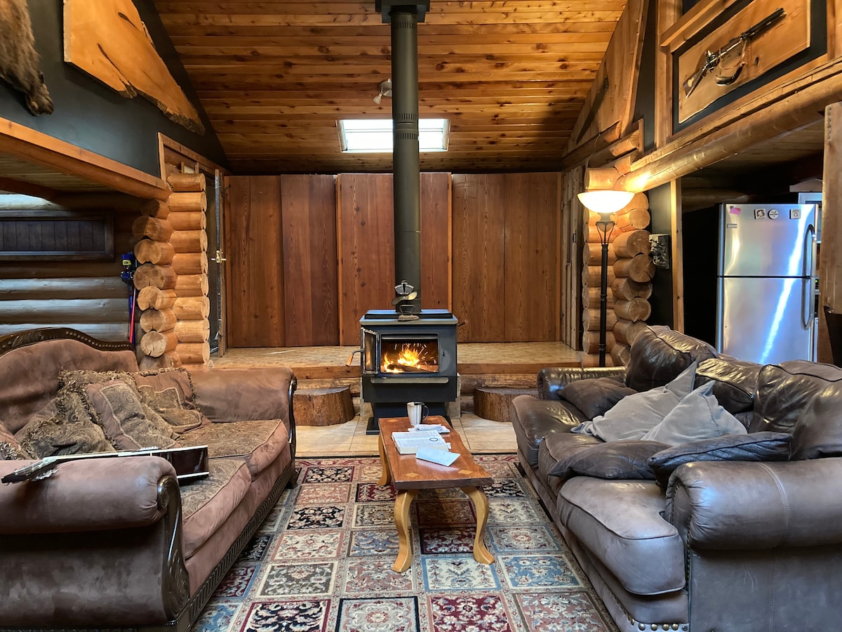 The living area features cozy leather sofas arranged around a rustic wooden coffee table. A wood stove provides warmth and ambiance, with logs stacked nearby. Natural wooden beams and a high ceiling enhance the cabin's rustic charm, while a patterned rug adds comfort.