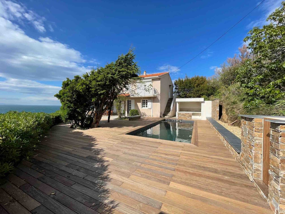 The outdoor space features a beautifully arranged wooden deck surrounding a rectangular pool. The house is visible in the background, set against a clear blue sky. Lush greenery frames the area, providing a natural and serene environment.