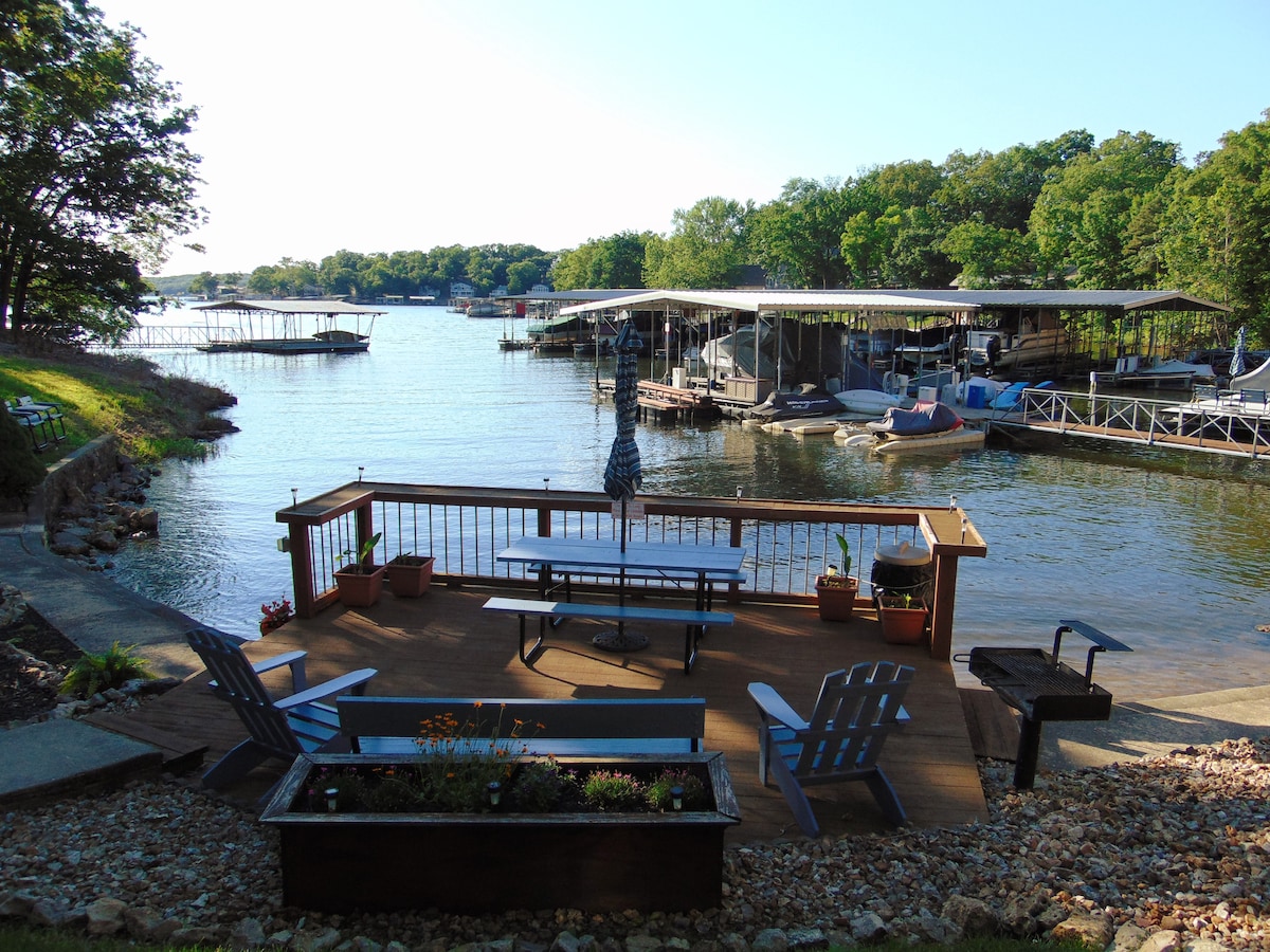 A scenic view of a lake area featuring a wooden deck with a picnic table and adirondack chairs. A small grill is positioned nearby. The lake is bordered by lush greenery, with docks visible in the background, providing space for various watercraft.