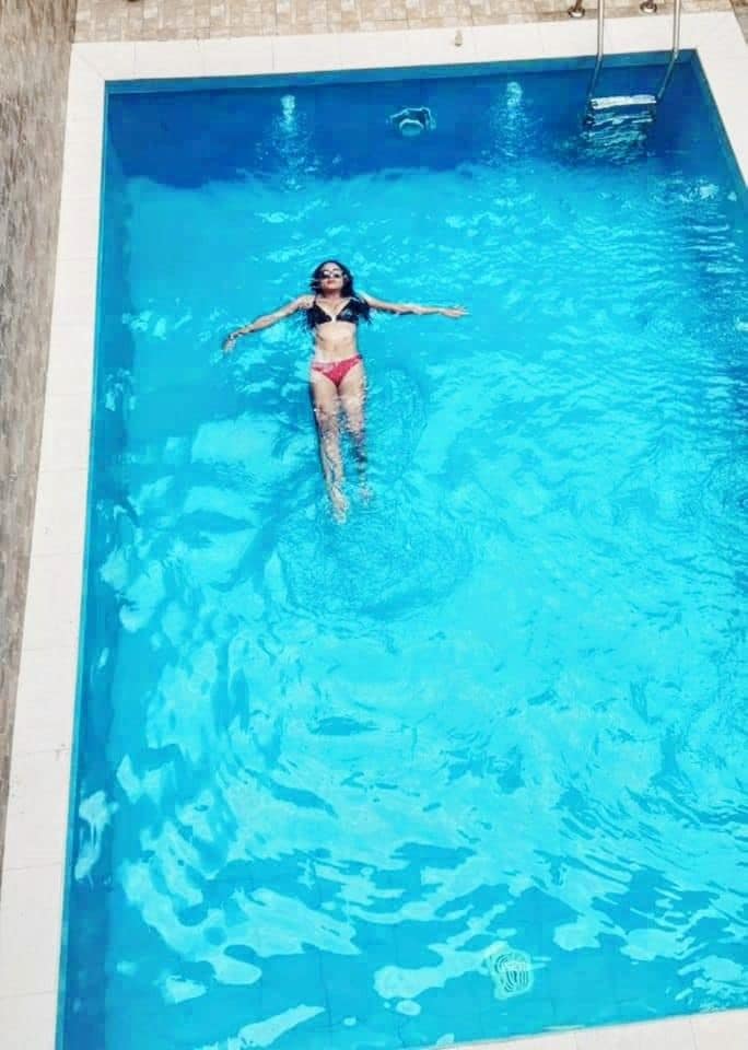 A clear blue swimming pool is depicted, featuring a person floating calmly on the water. The pool's tiles create a neat border, and a ladder is visible on one side, allowing for easy access. Sunlight reflects off the water surface, enhancing the tranquil environment.