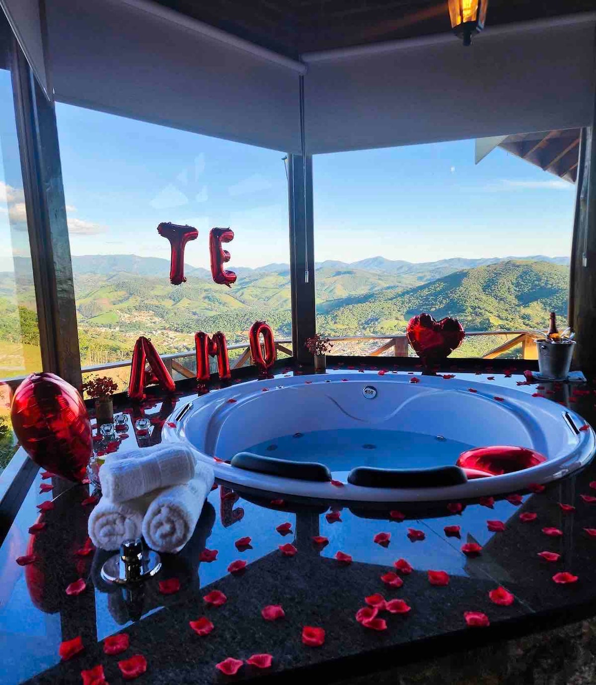 A jacuzzi sits adjacent to large windows, offering a panoramic view of rolling mountains. The space is adorned with red rose petals and balloons spelling 'TE AMO.' White towels are neatly arranged beside the tub, enhancing the romantic setting.