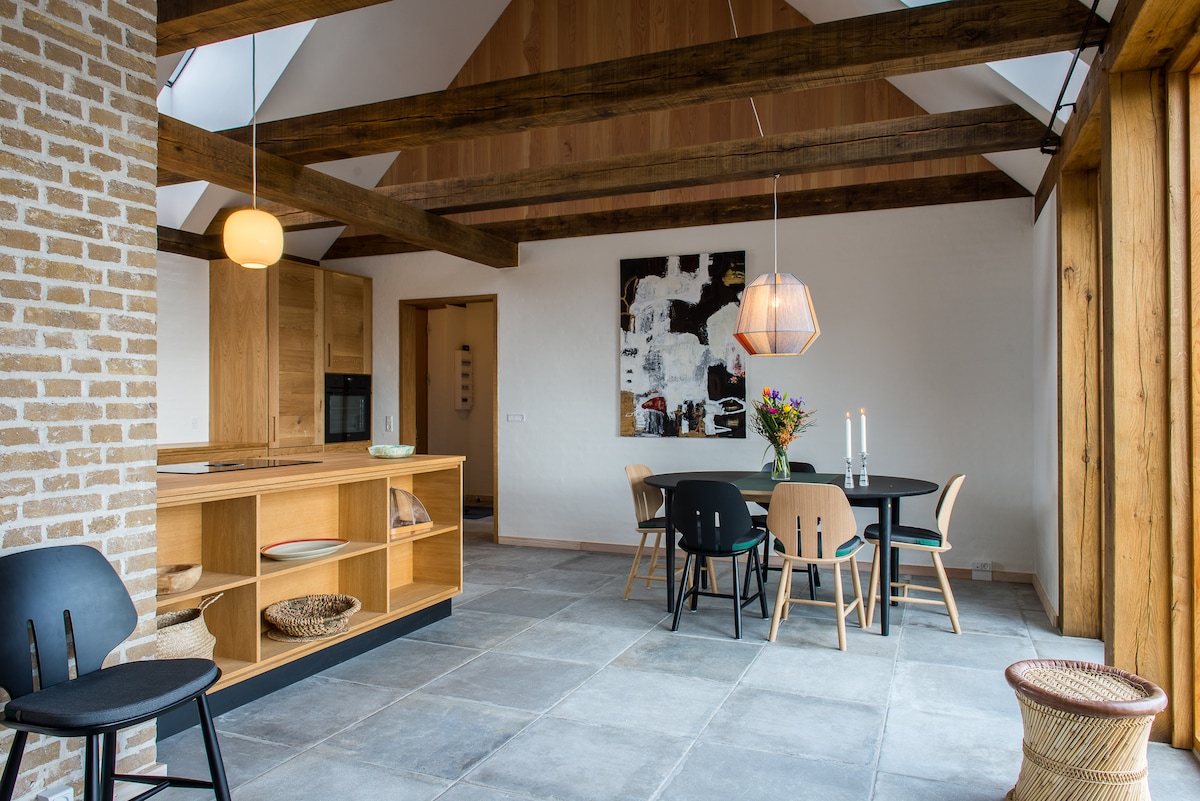 An open dining area features a round table surrounded by six chairs, set against a backdrop of exposed wooden beams and brick walls. Natural light fills the space through large windows, highlighting a minimalist décor complemented by a modern pendant light and fresh flowers.
