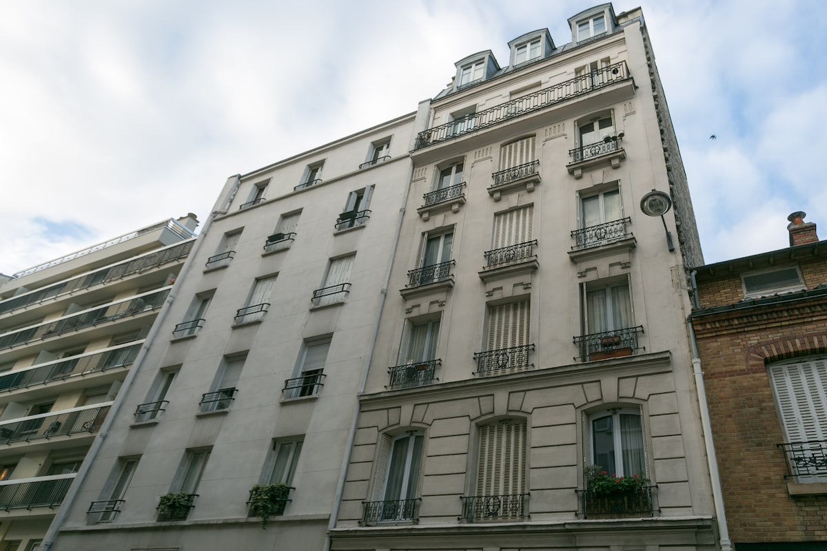A classic Parisian building stands elegantly, showcasing its decorative iron balconies and cream-colored façade. The structure features distinct architectural elements, with several large windows framed by ornamental detailing. The clouds above suggest a calm atmosphere in the surrounding urban environment.