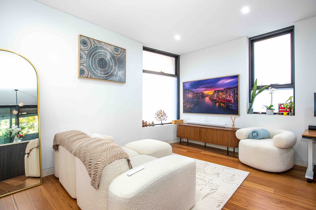 A stylish living area features a plush, light-colored sofa and soft armchairs, complemented by a wooden media unit. Large windows provide natural light, while a colorful artwork hangs above the TV. A cozy blanket is draped over the sofa, enhancing the inviting atmosphere.