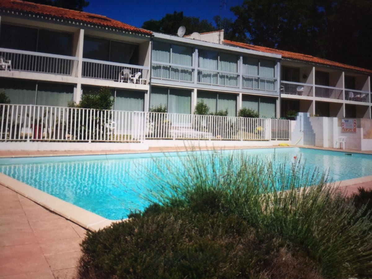 A private residence is depicted with a clear blue swimming pool in the foreground. The building features multiple balconies with white railings, surrounded by green foliage. Sunlight reflects off the water, creating a serene environment.