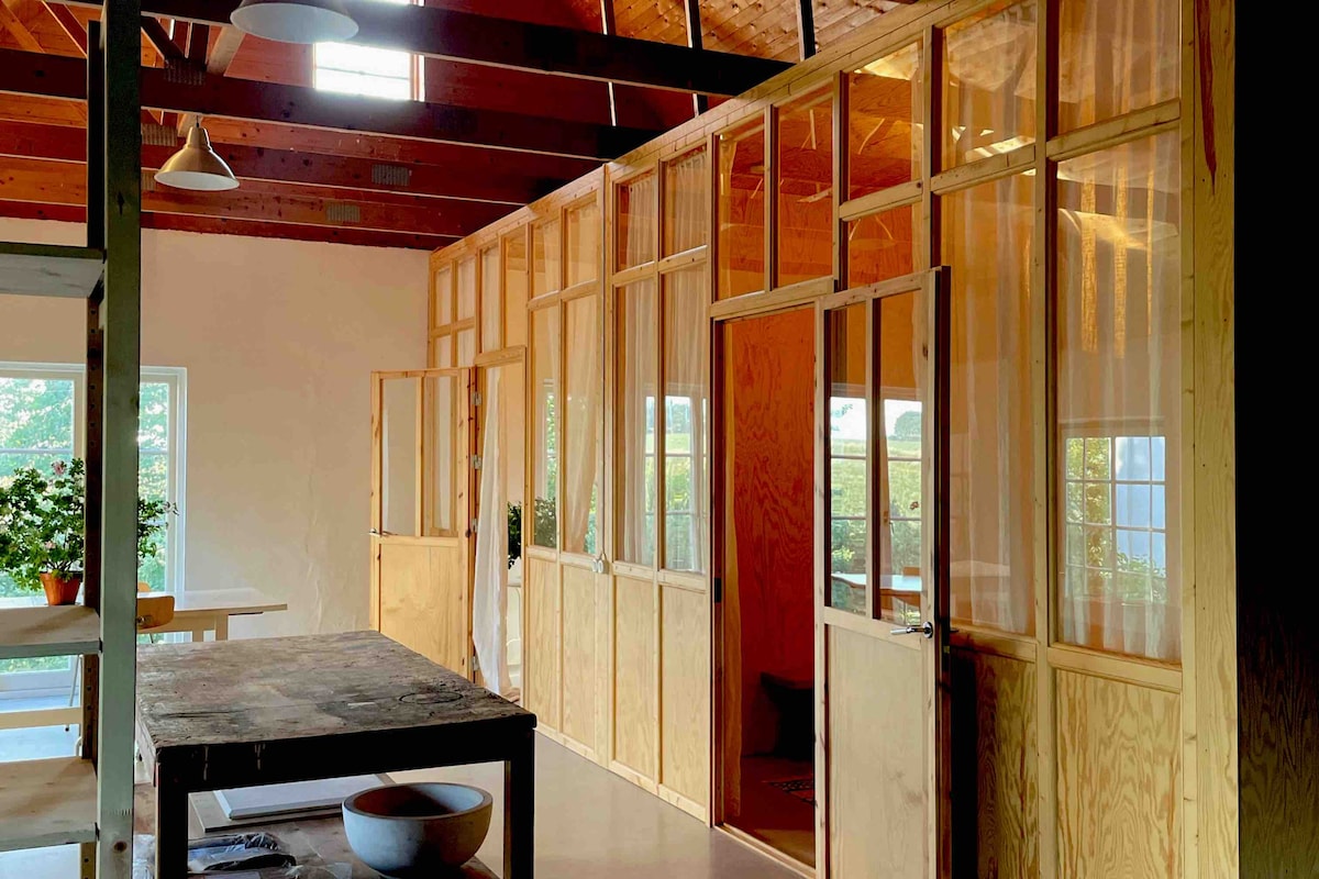 A spacious area features wooden partitions with large windows and curtains, allowing natural light to fill the room. A long table is positioned in the foreground, surrounded by various plants. The design reflects a contemporary, open space atmosphere.