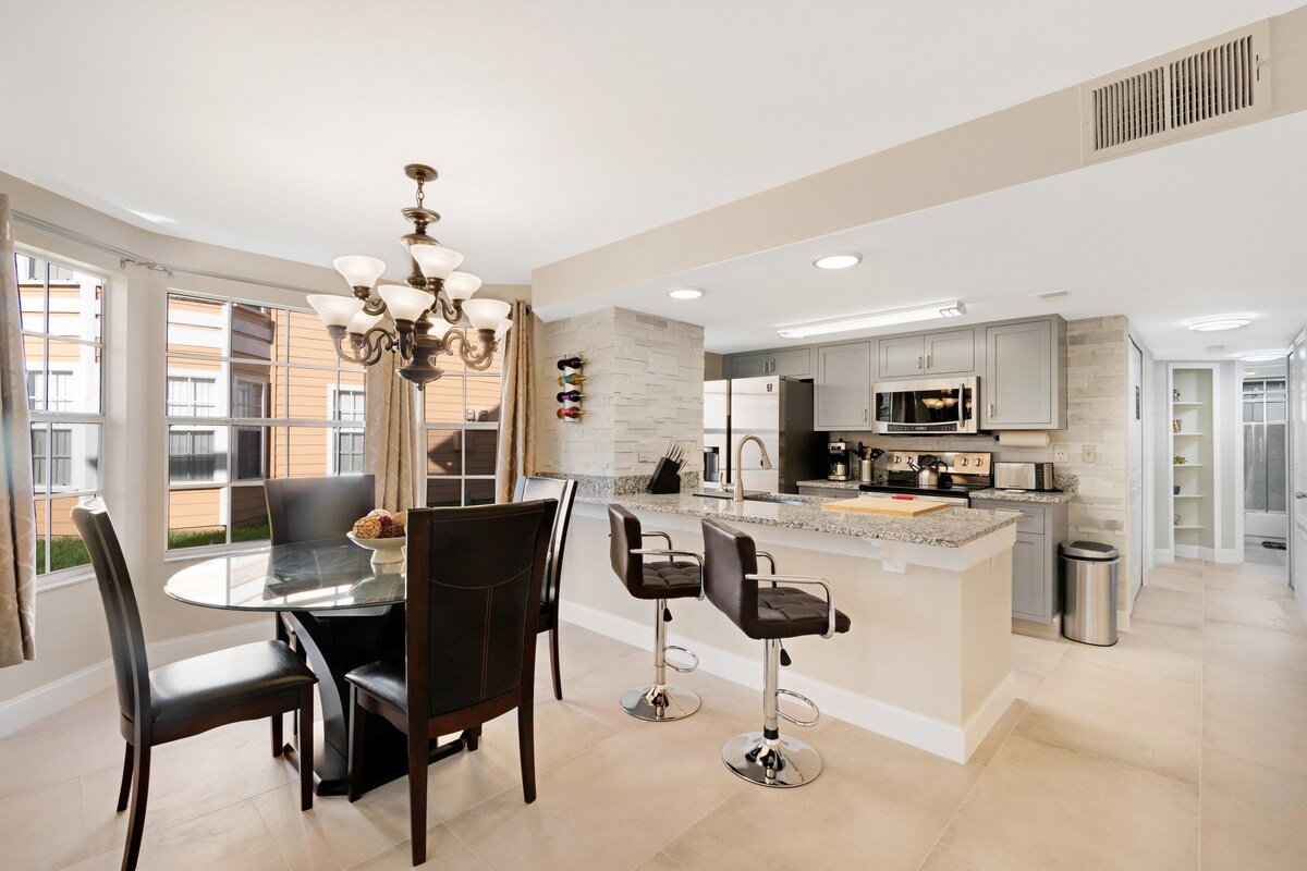 A modern kitchen and dining area showcases a round glass table surrounded by four black chairs. Bar stools are positioned at the kitchen counter, and stainless steel appliances are integrated into the cabinetry. Large windows provide natural light, enhancing the open and airy feel of the space.