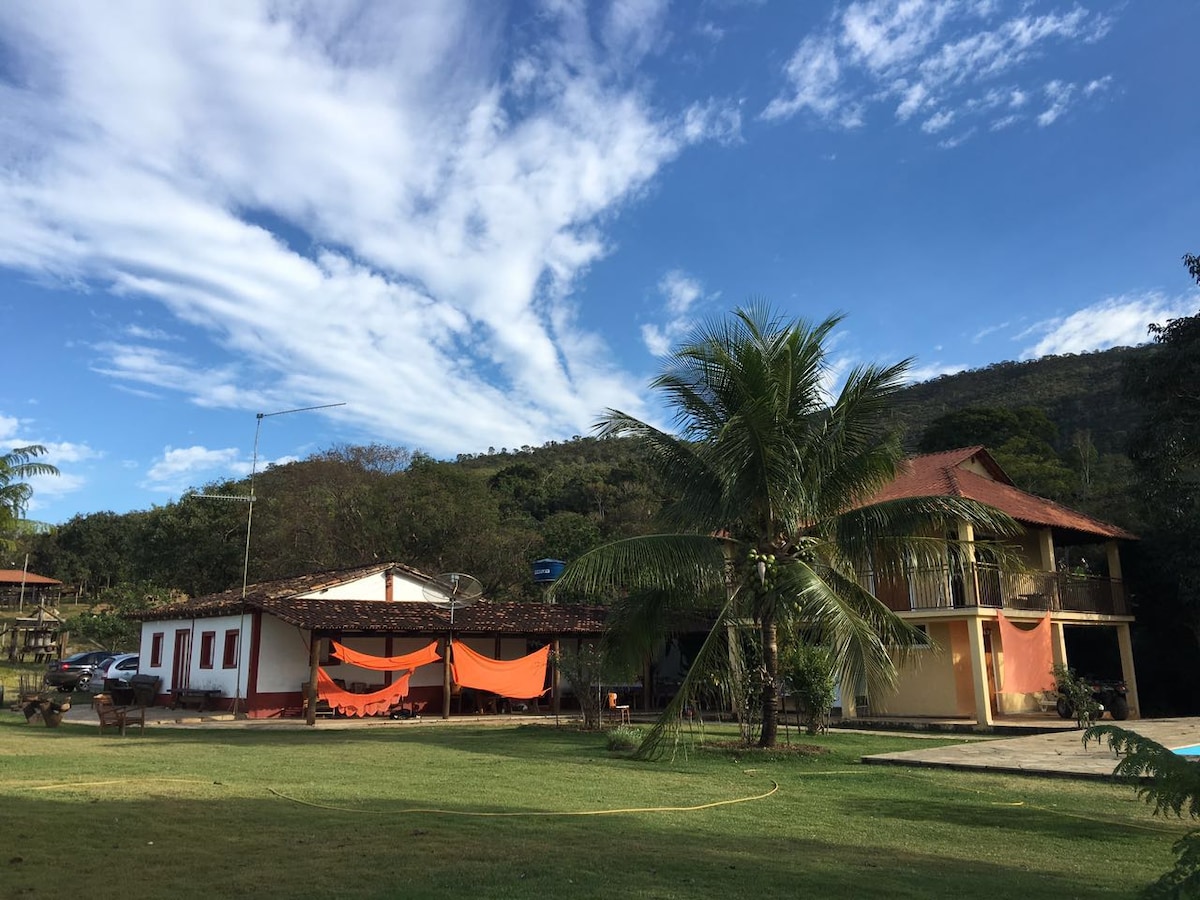 A spacious outdoor area showcases two structures with varying architecture, surrounded by lush greenery and a blue sky. Hammocks are suspended under colorful fabric canopies, providing shaded relaxation spots. A clear pool area is visible, reflecting the surrounding landscape.