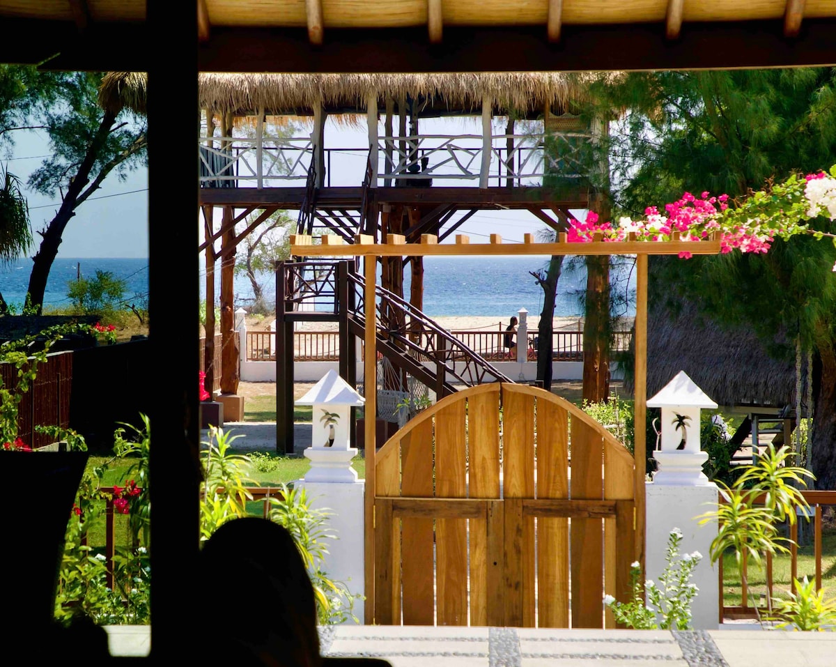 A wooden gate opens to a path leading to the beachfront, framed by tropical plants and a distant view of the ocean. A multi-level structure with a thatched roof is visible, enhancing the outdoor space's inviting atmosphere.