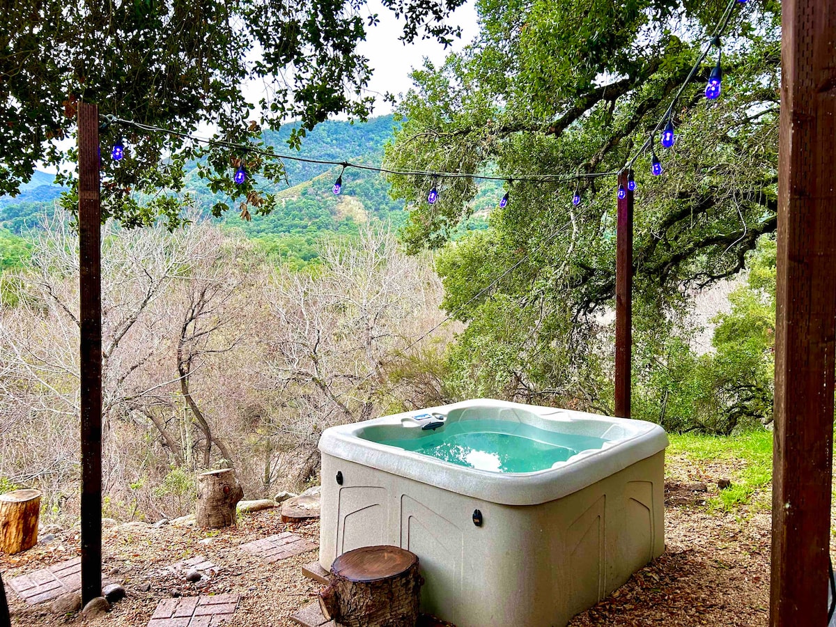 A hot tub is surrounded by natural scenery, framed by wooden posts adorned with blue string lights. Leafy trees provide shade, and the landscape features rolling hills in the background, adding to the serene atmosphere of the outdoor space.