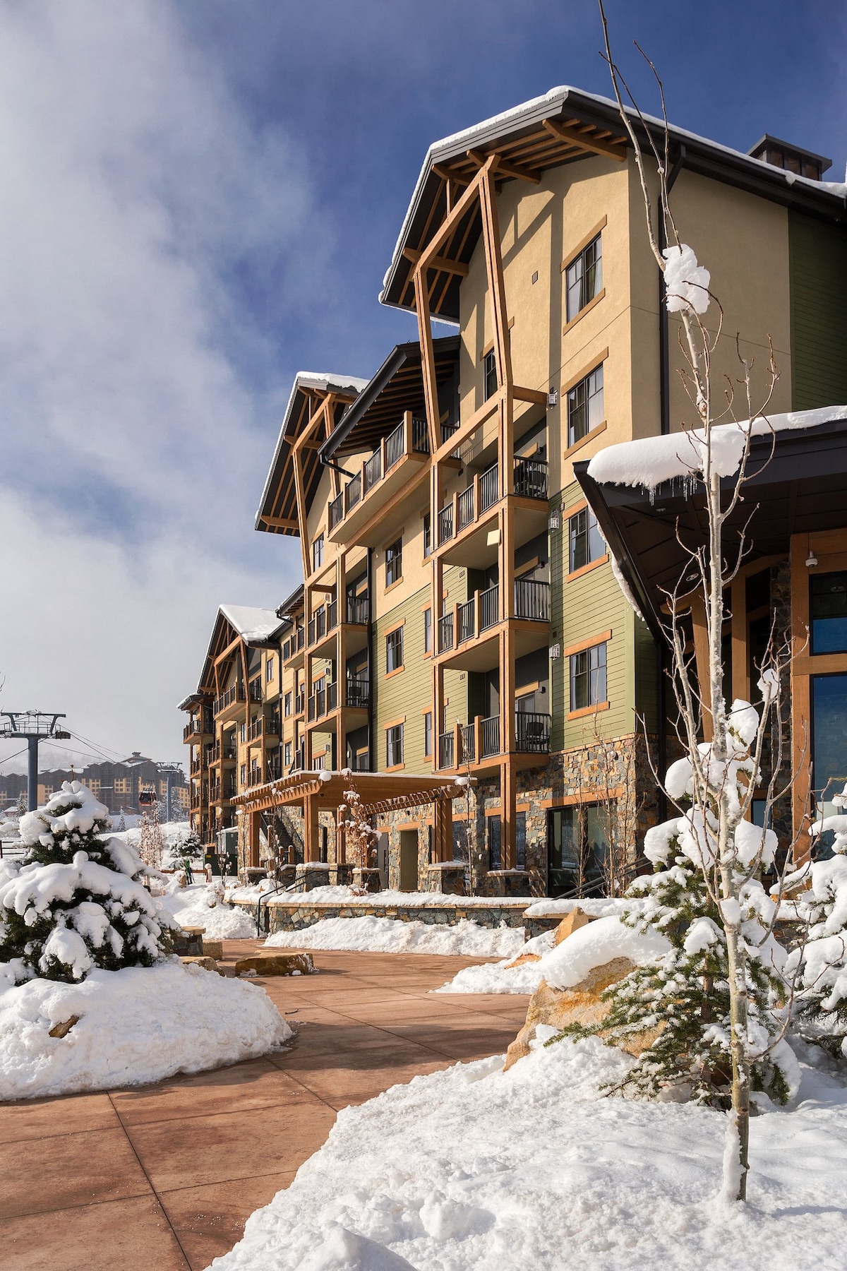 The exterior of the resort is shown, featuring a multi-story building adorned with large windows and wooden accents. Snow blankets the surrounding landscape, while snow-covered trees line the pathway leading to the entrance. A gondola lift is visible in the background, connecting to the ski slopes.