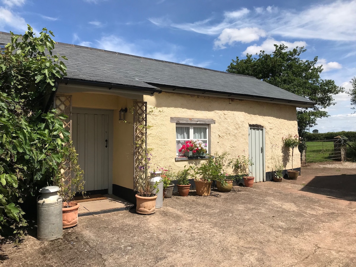 The entrance to the Little Barn is framed by potted plants and flowers, adding a touch of color to the exterior. The building features a light yellow facade and a slate roof, with a wooden door and window offering a glimpse of the inviting interior.