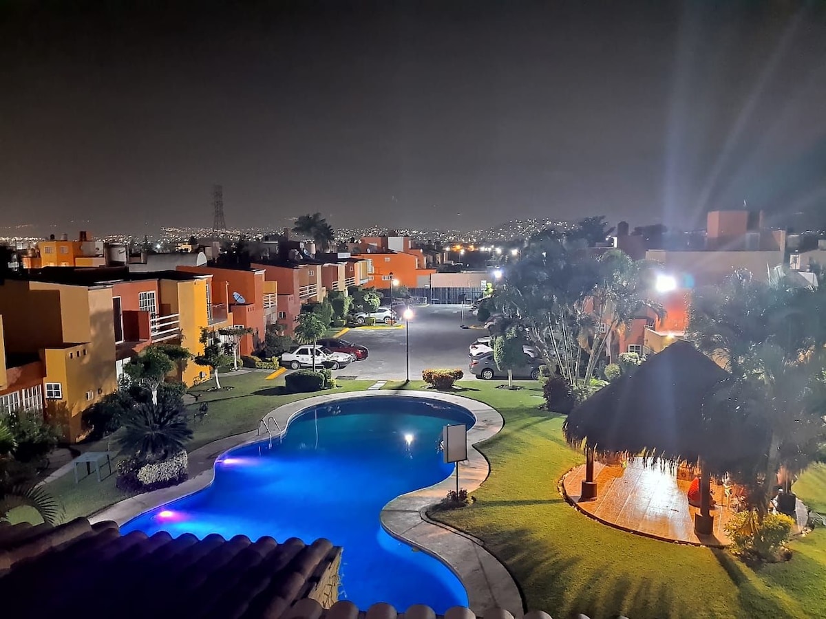 A well-lit community is depicted at night, featuring a curvilinear pool reflecting blue lights. Surrounding the pool are landscaped green areas, palm trees, and a thatched-roof palapa. Nearby, several houses and parking spaces are visible, enhancing the inviting atmosphere of the environment.