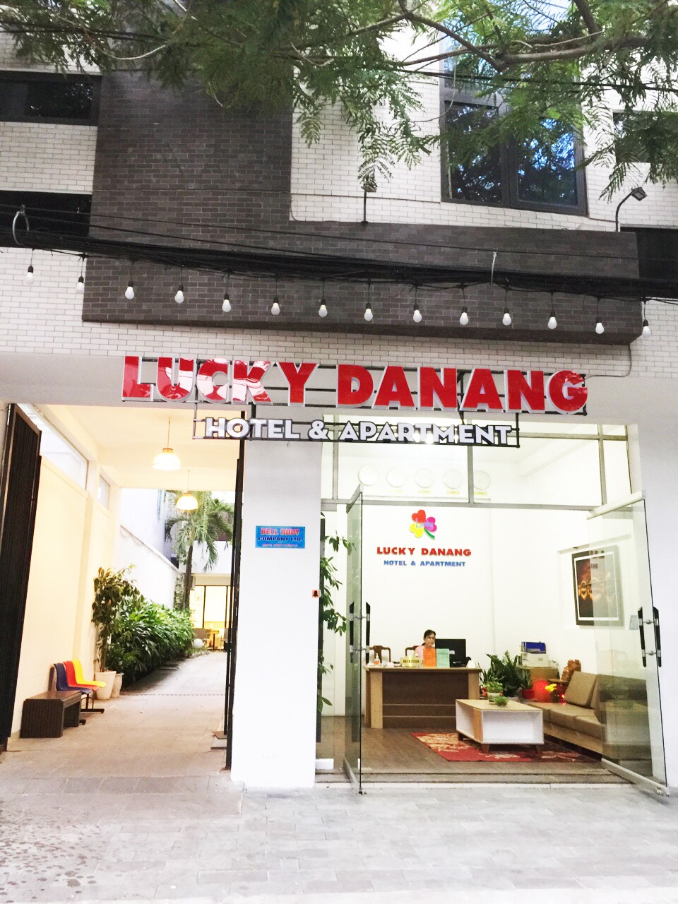 The entrance of the Lucky Danang Hotel & Apartment is visible, framed by lush greenery. A welcoming reception area is showcased through large glass doors, featuring modern decor with seating arrangements. The pathway leads into the hotel, providing a sense of accessibility and openness.