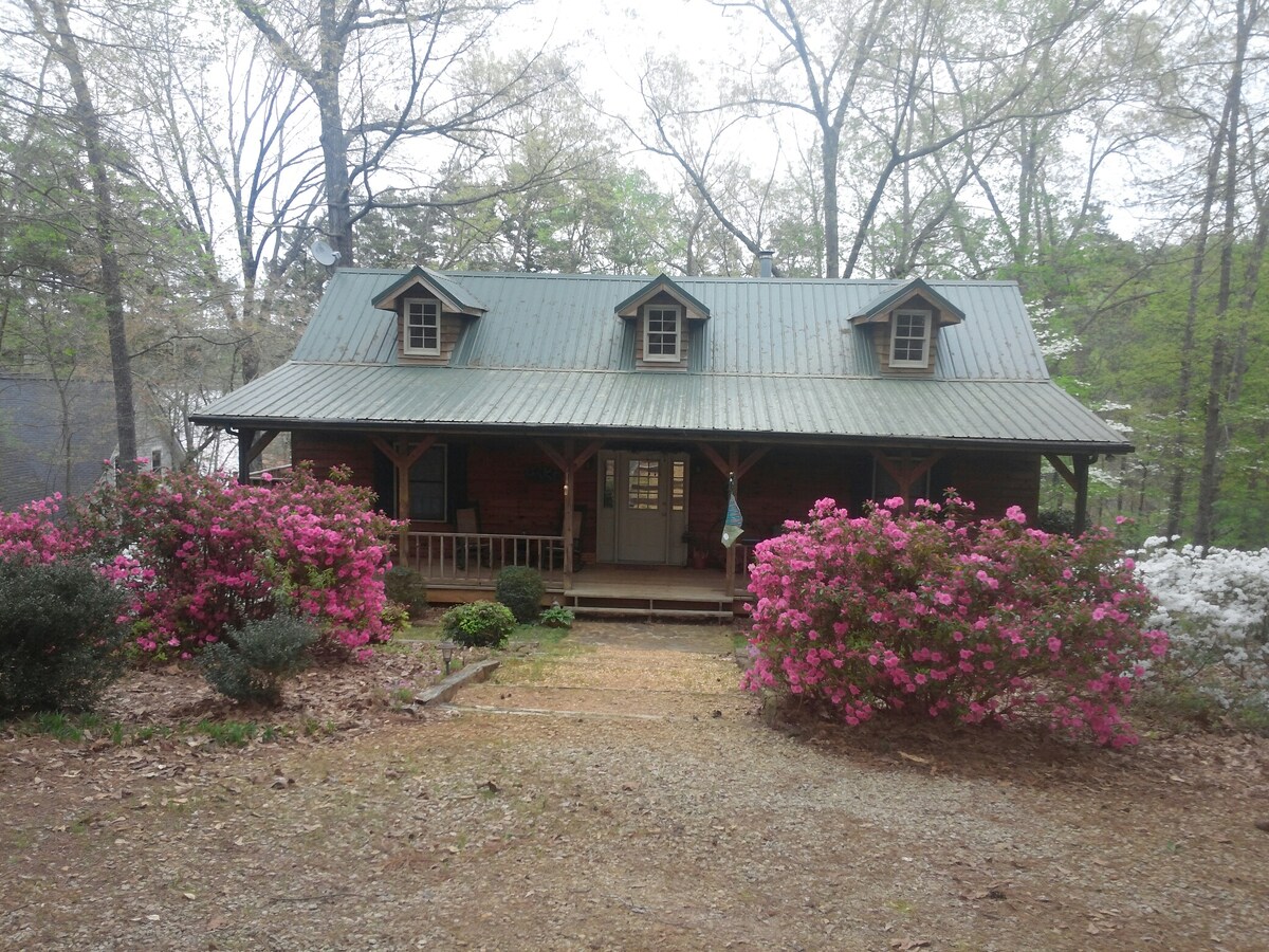 The exterior of the Cape Cod style cabin features a metal roof and charming dormer windows. Vibrant azaleas in pink and white adorn the front yard, complemented by gravel pathways leading to a welcoming front porch with seating.