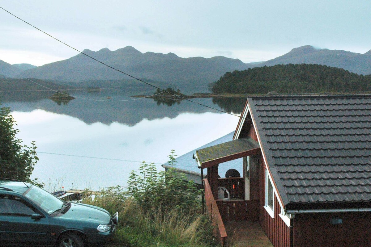 A holiday house is situated on a hillside, overlooking a tranquil fjord. Dark mountains are visible across the water, with a few scattered trees lining the shore. A car is parked near the entrance, and the exterior features traditional wooden siding.