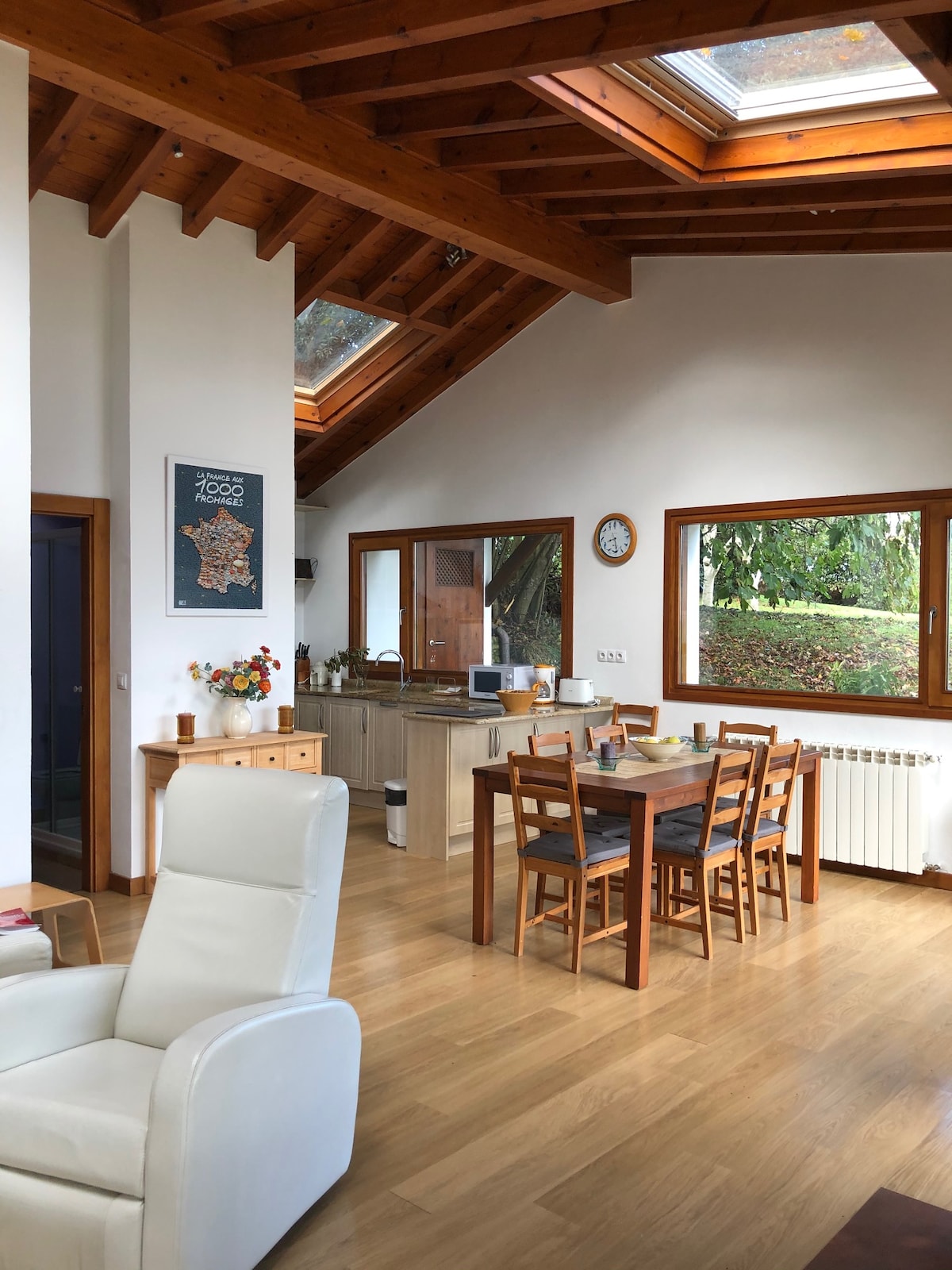 An open living space features a dining area with a wooden table surrounded by chairs. Skylights and large windows allow natural light to illuminate the room. A cozy sitting area is designated with a white chair, while a kitchen equipped with modern appliances is visible in the background.