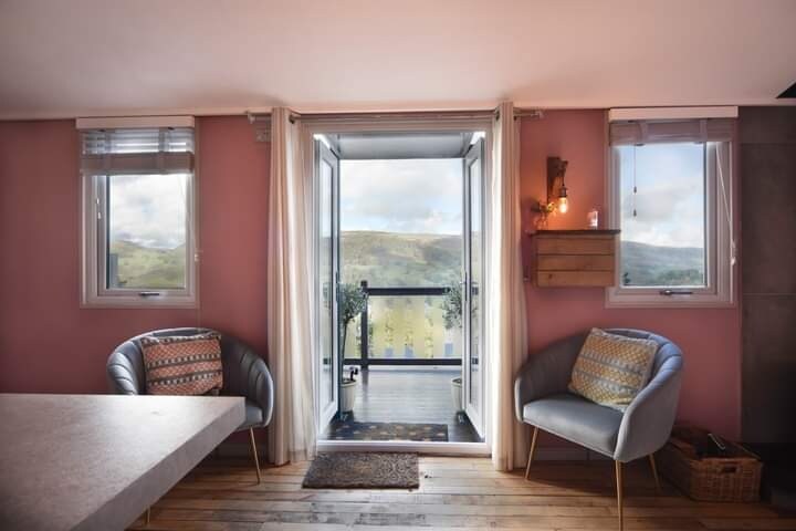 A cozy interior space features two soft armchairs, each adorned with decorative cushions. Natural light floods in through double glass doors that open to a scenic view of the hillside. The wooden floor and light pink walls create a warm, inviting ambiance.