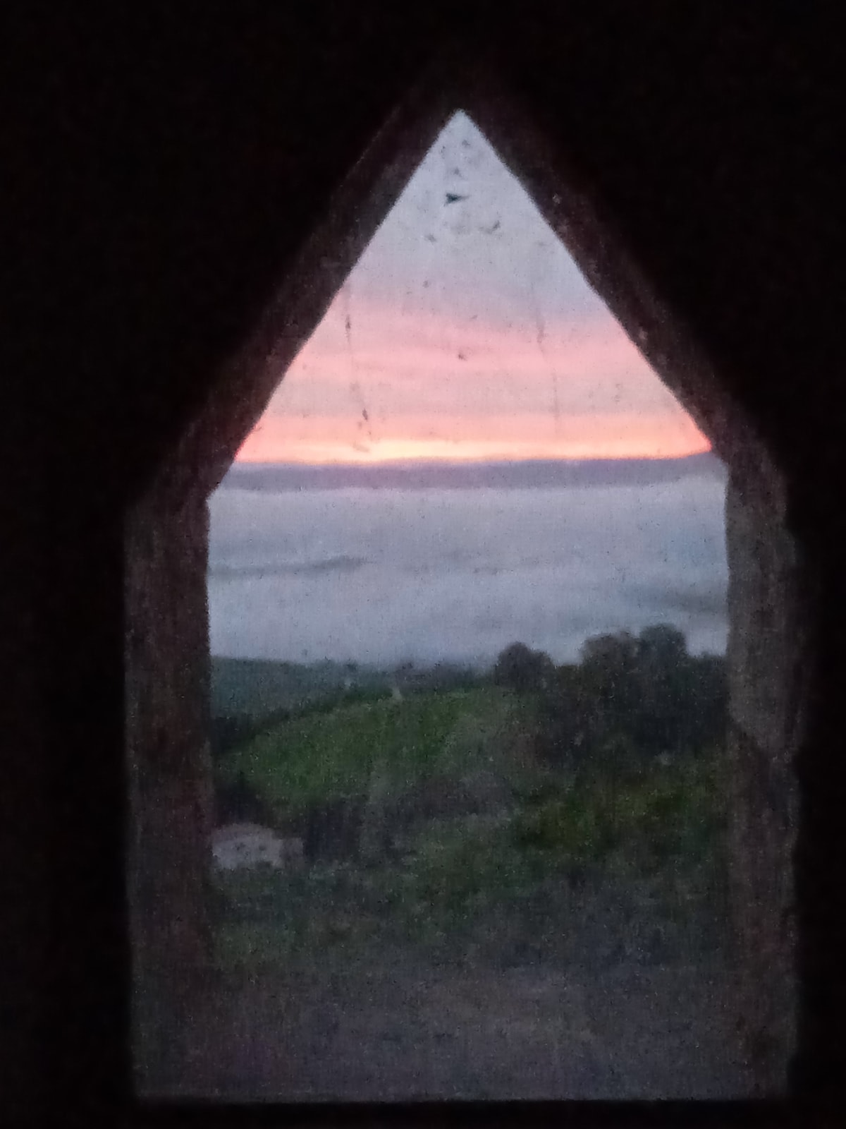 A triangular window frames a serene view of a sunrise over rolling hills. Soft colors of pink and orange illuminate the sky while fog blankets the landscape below, creating a peaceful and tranquil atmosphere.