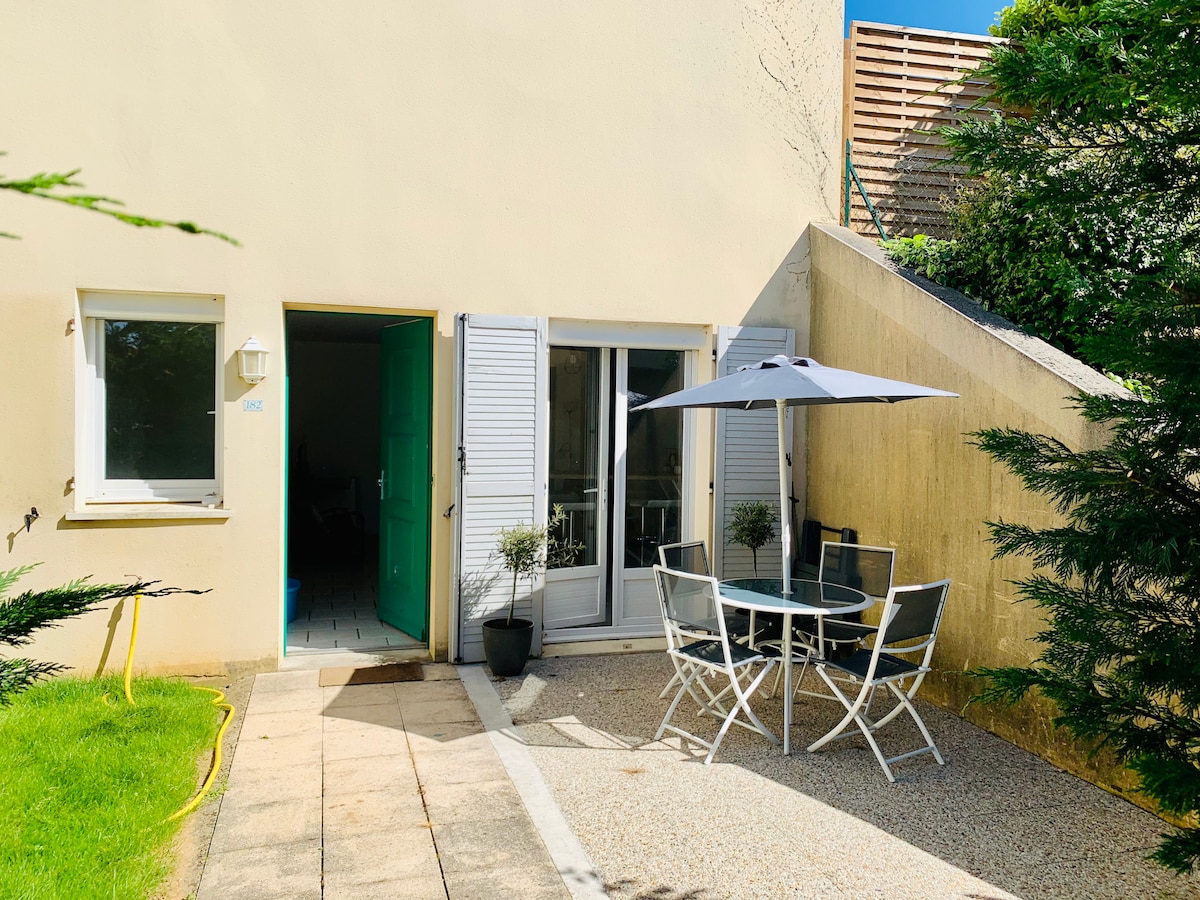 A private terrace is presented, furnished with a table and four chairs, shaded by a parasol. Green grass borders the area, with a clear pathway leading to the entrance. Nearby, two windows are visible, allowing natural light into the space.